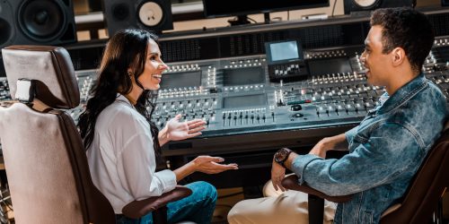 two smiling multicultural sound producers sitting by mixing console in recording studio